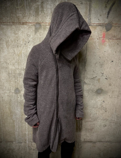 Person wearing a long, dark gray hooded coat against a textured wall.