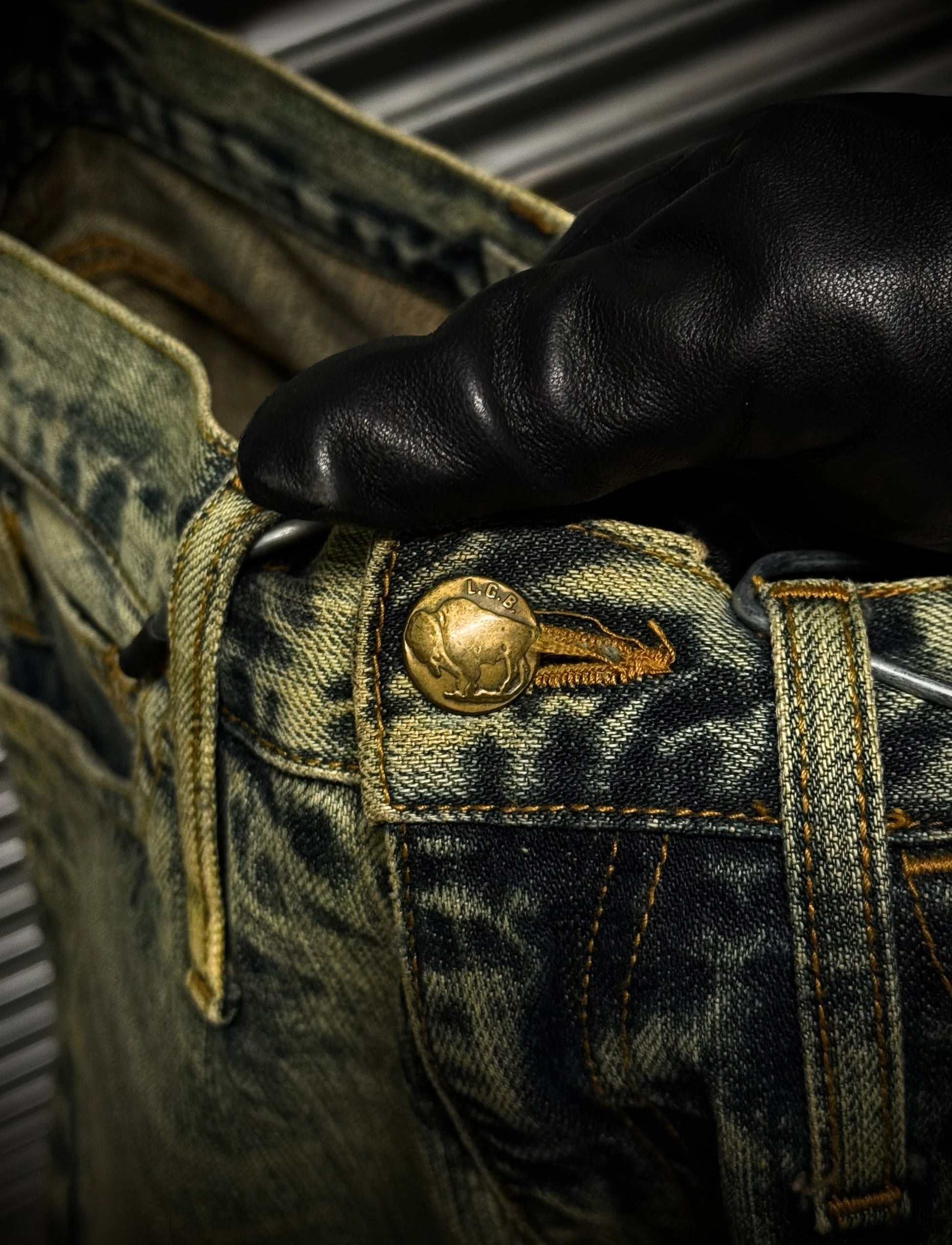 Close-up of a hand wearing a black glove touching a gold button on a pair of green denim jeans.