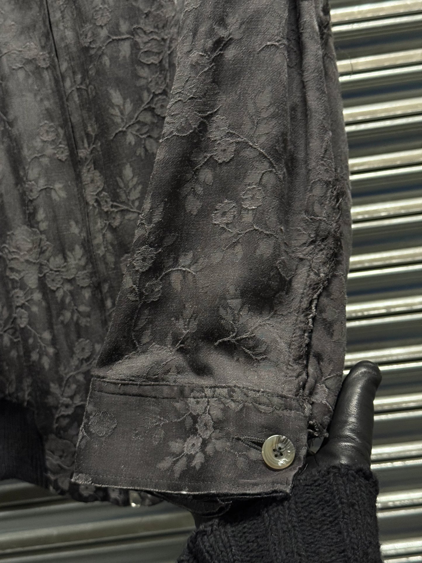 Dark floral-patterned jacket with a button, held by a gloved hand against a metallic background.