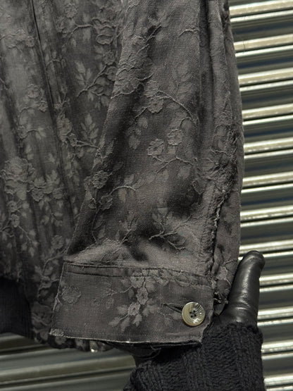 Dark floral-patterned jacket with a button, held by a gloved hand against a metallic background.