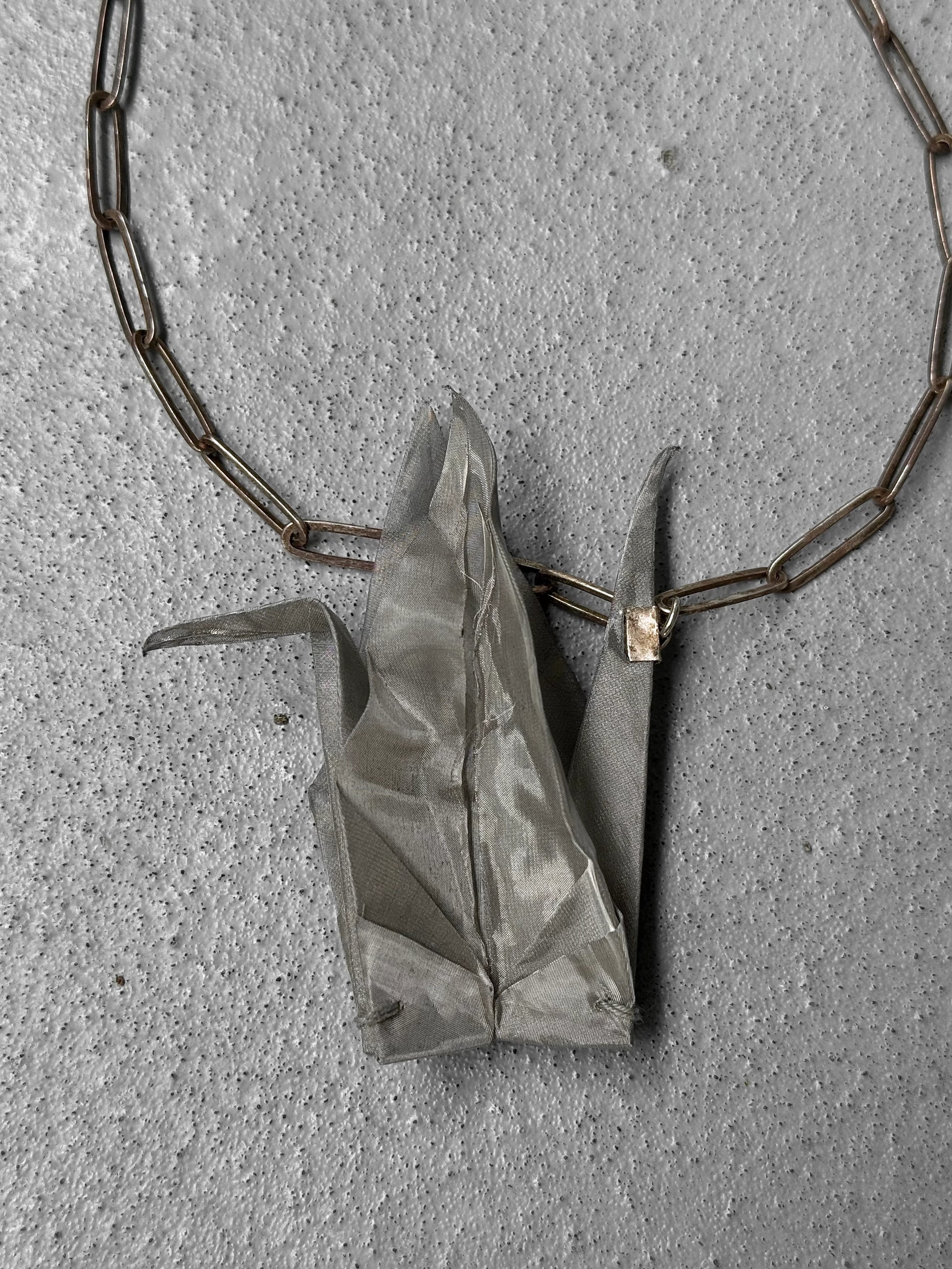 Silver origami crane pendant on a chain against a gray concrete background